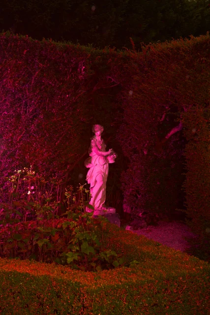 Statue of a woman holding a basket stands in a garden with green plants and a sculpted hedge under red and pink lighting.