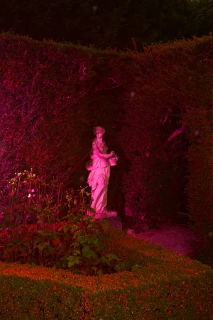 Statue of a woman holding a basket stands in a garden with green plants and a sculpted hedge under red and pink lighting.