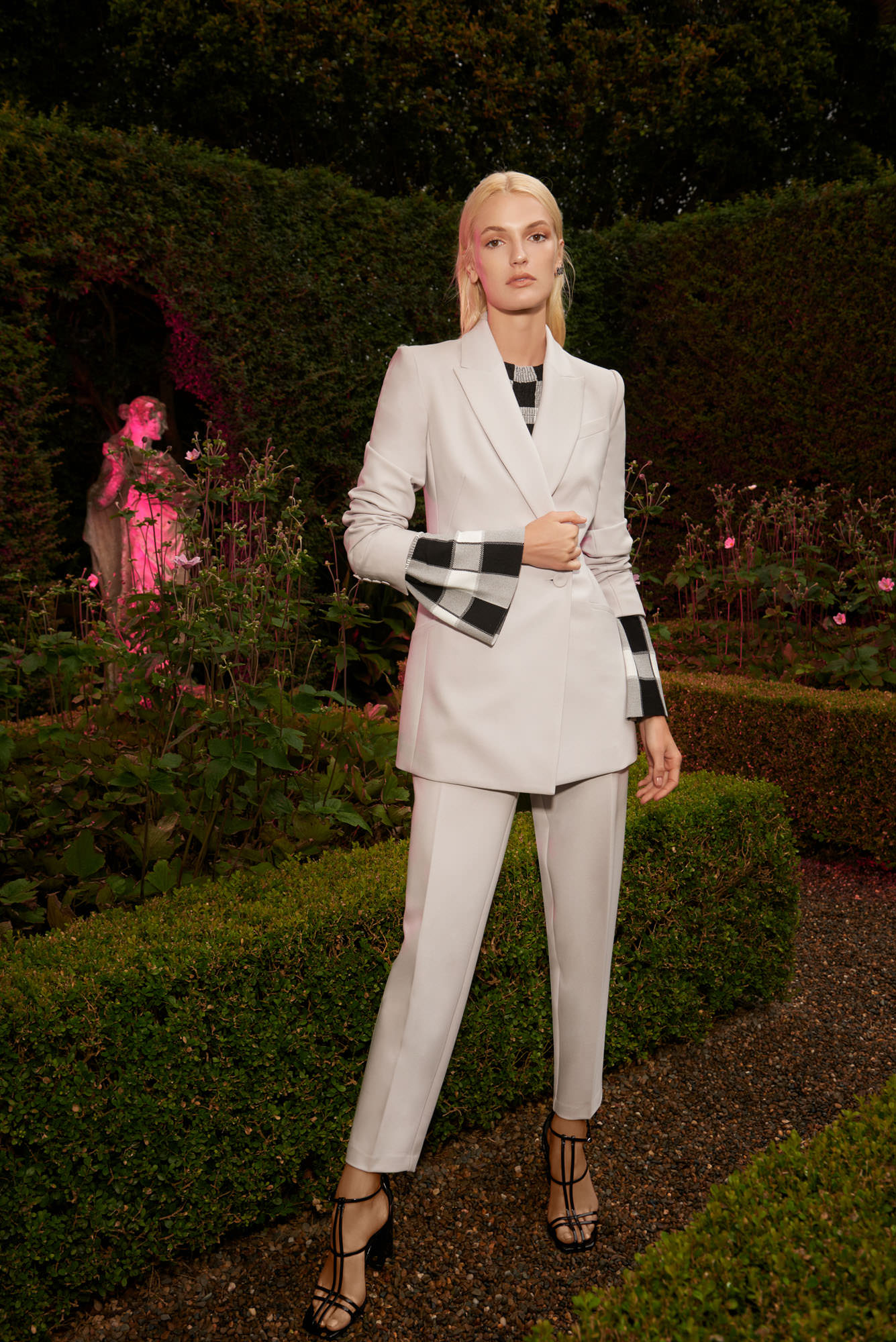 Woman in a grey blazer and pants suit stands in a garden setting with trimmed hedges and a statue in the background.