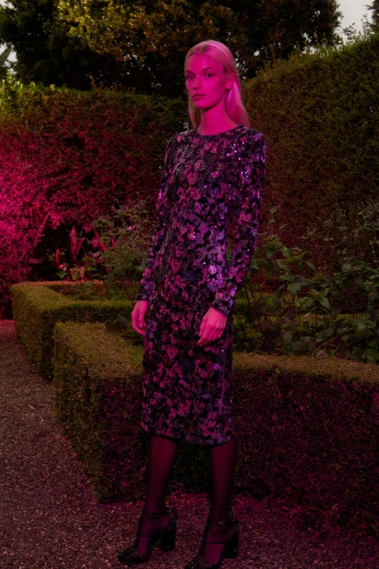 Full shot of a blonde adult woman in a long-sleeved floral midi dress and heeled sandals, posing in a garden against a foliage background.