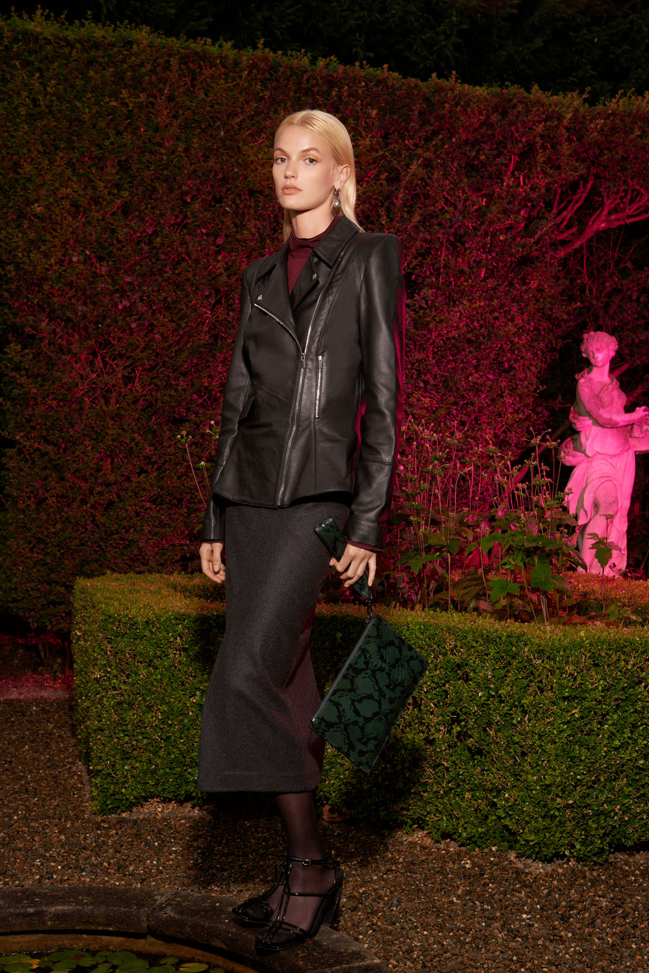 A fair-skinned adult woman models a black leather jacket, dark skirt, and patterned clutch in an outdoor garden setting with pink lighting.