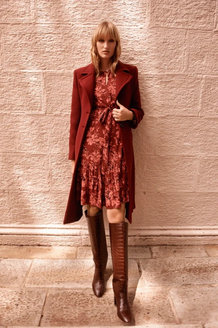 Adult female fashion model poses in floral dress, long coat, and knee-high crocodile-skin boots against a stone wall.