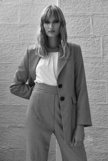 Woman in black and white checkered suit stands against a textured wall, showcasing high-waisted pants and a buttoned blazer, indoors.