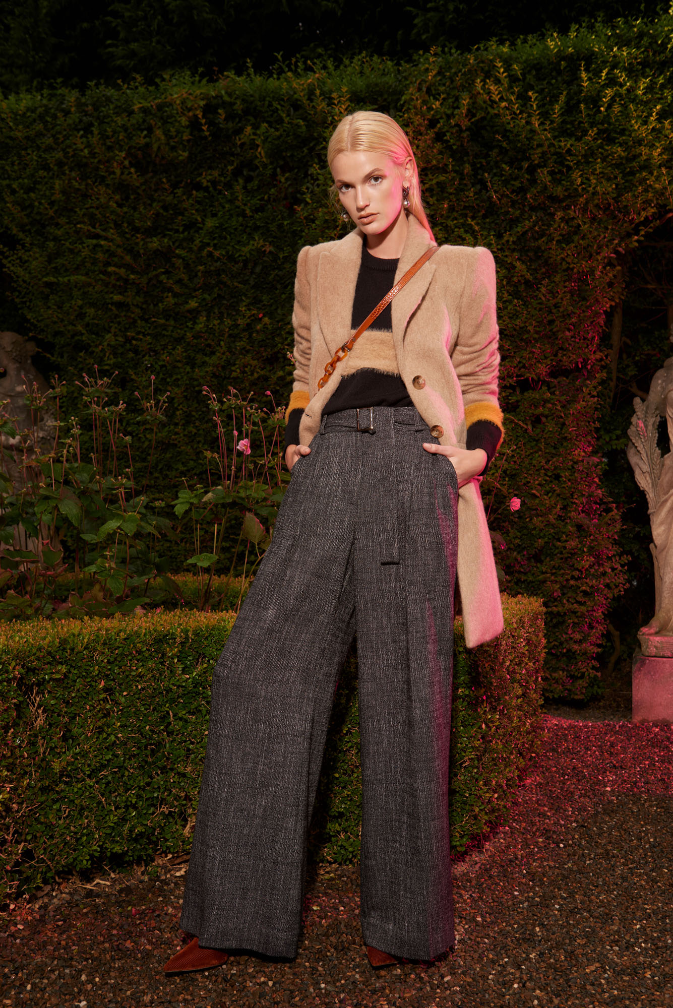 Fashionable adult female model poses outdoors in a tan jacket, black top, and grey wide-leg trousers with green hedge background.