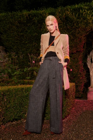 Model with blonde hair wearing a beige blazer and grey wide-leg trousers standing outdoors before hedges.