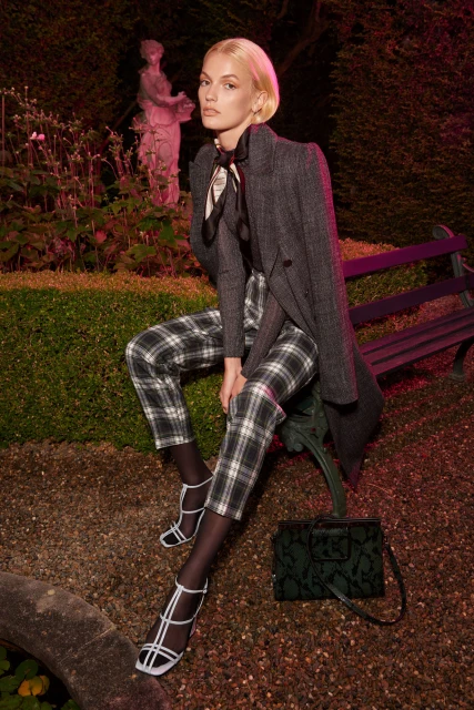 A fair-skinned woman with blonde hair sits on a park bench, wearing a gray tweed coat, plaid pants, white heels, and a snakeskin purse.