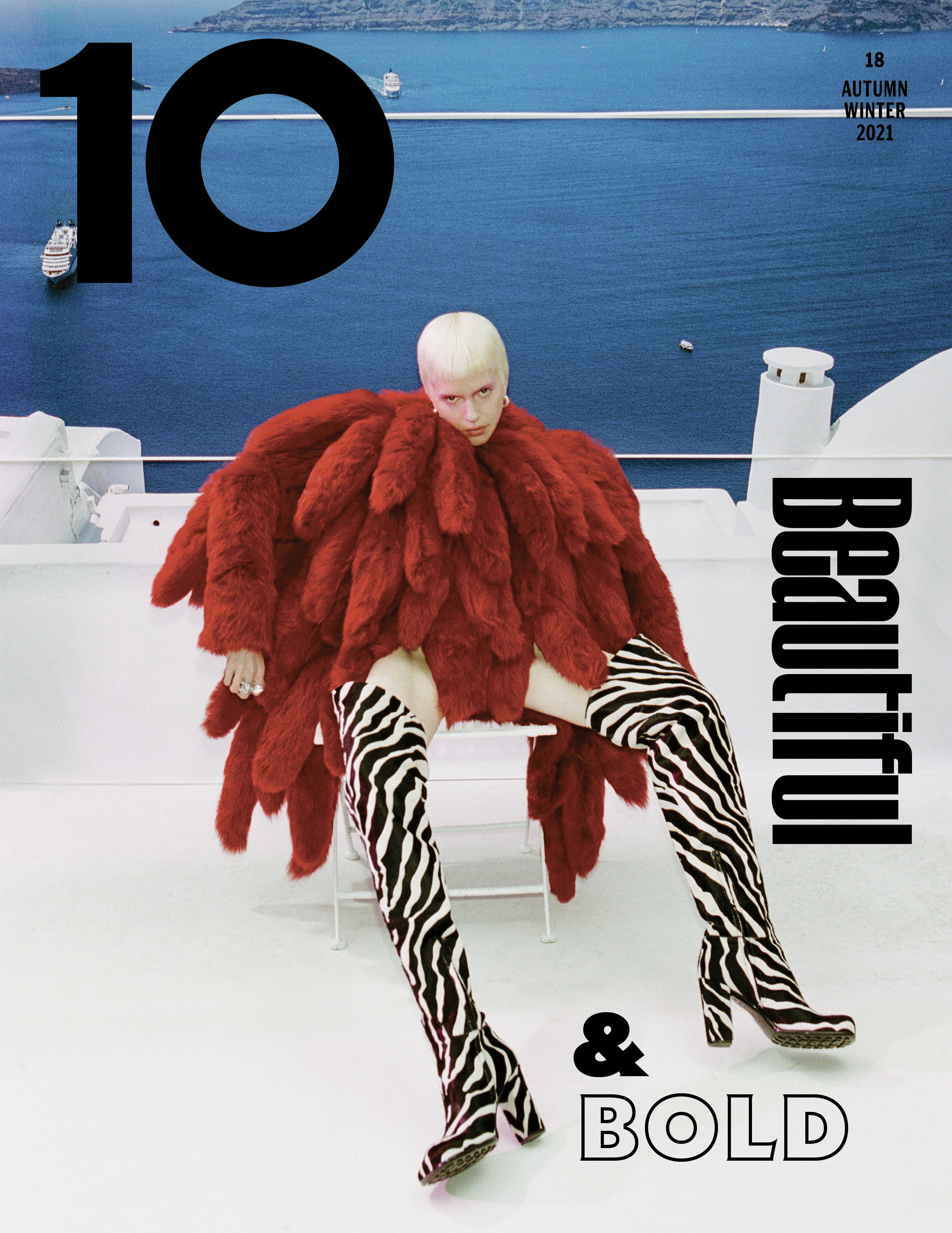 Magazine cover featuring a model in red fur-like coat and zebra-striped boots, posing against a blue ocean backdrop with white architecture.