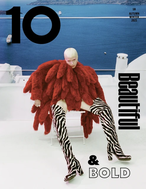 Magazine cover featuring a model in red fur-like coat and zebra-striped boots, posing against a blue ocean backdrop with white architecture.