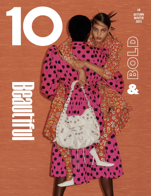 Two fashion models posing in patterned dresses and white high heels for a magazine cover, set against a textured orange background.