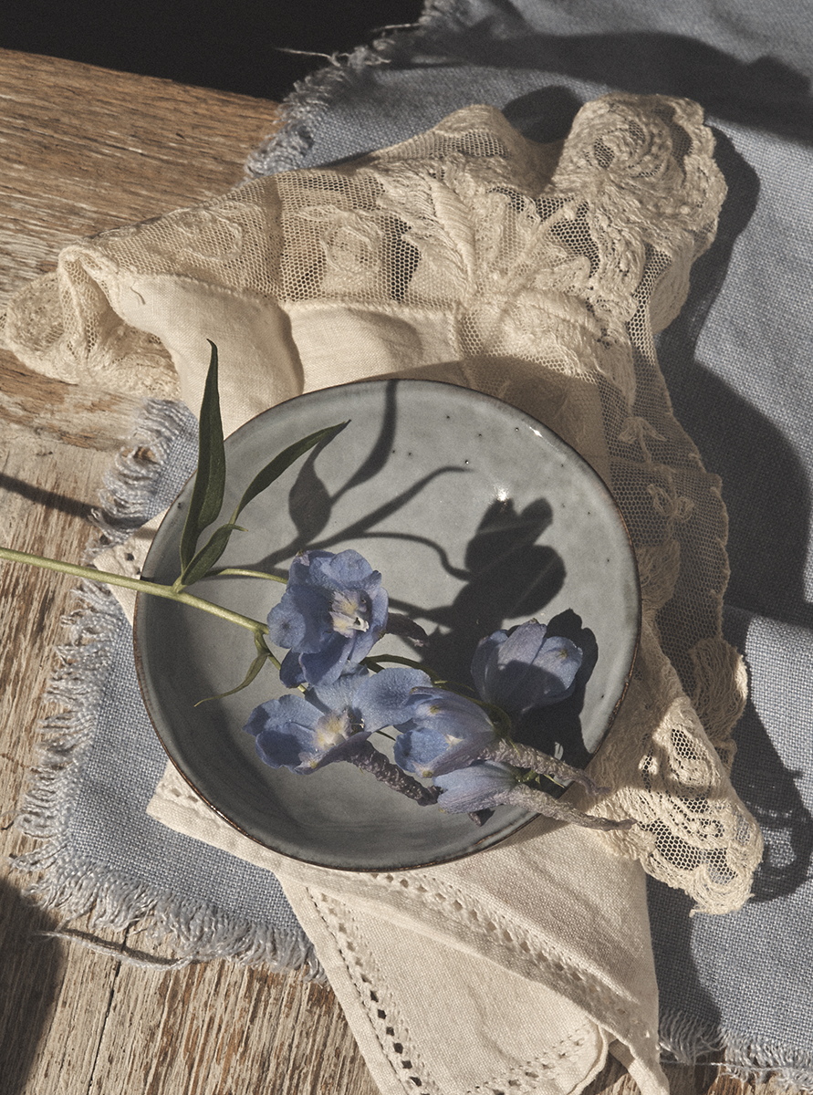 Overhead shot of a ceramic plate holding blue flowers on top of wooden table with linen fabric, capturing natural sunlight.