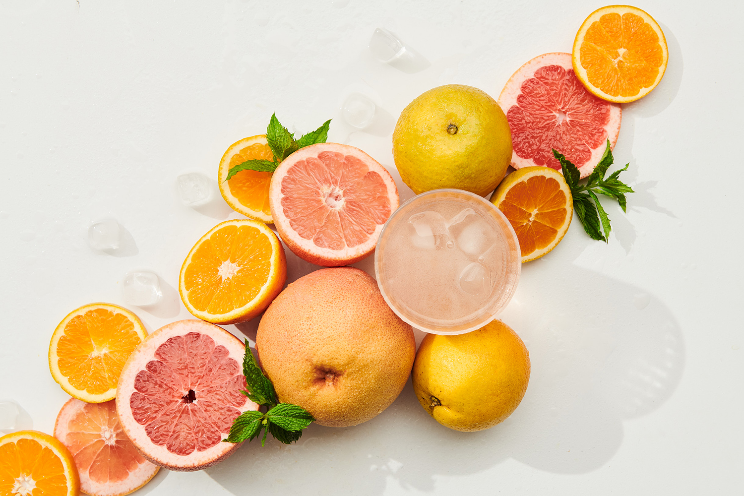 Overhead shot of citrus fruit assortment featuring oranges, grapefruits, lemons, a drink with ice cubes, and mint leaves against a white background.