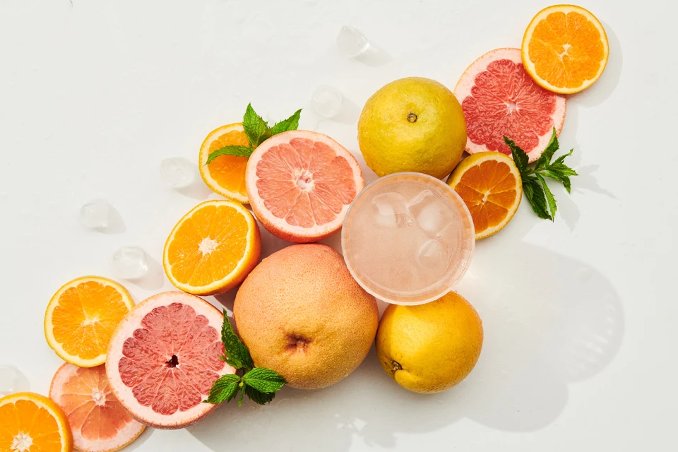 Overhead shot of citrus fruit assortment featuring oranges, grapefruits, lemons, a drink with ice cubes, and mint leaves against a white background.