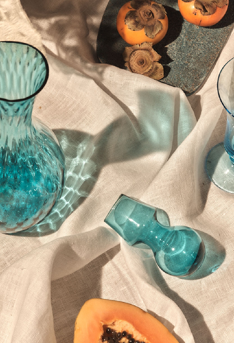 Still life shot of a turquoise glass vase, drinking glass, and small pipe with persimmons and papaya on a white linen cloth.