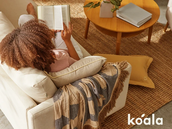 Woman with curly hair relaxes on a beige couch in a sunlit room reading a book, with a throw blanket, pillow, and a round wooden table nearby.
