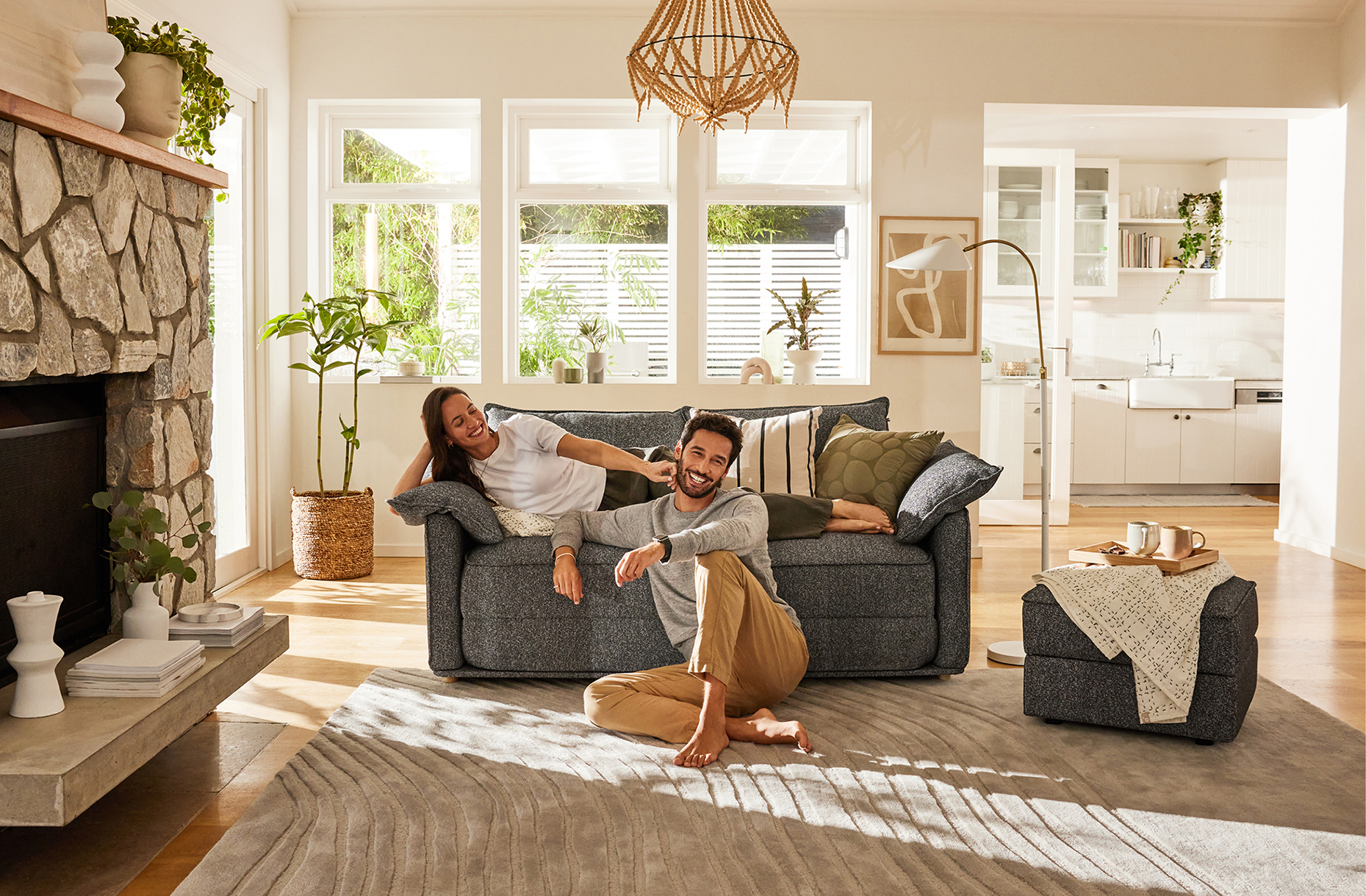 A couple relaxes on a gray sofa in a naturally lit living room with stone fireplace, wood floors, and neutral-toned decor and furnishings.