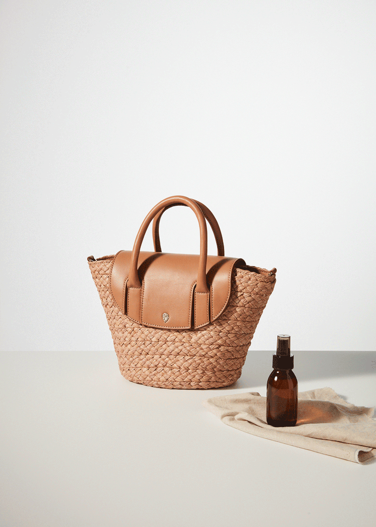 A straw tote bag with leather flap and handles sits on a light surface next to a brown spray bottle and cleaning cloth in a product shot.