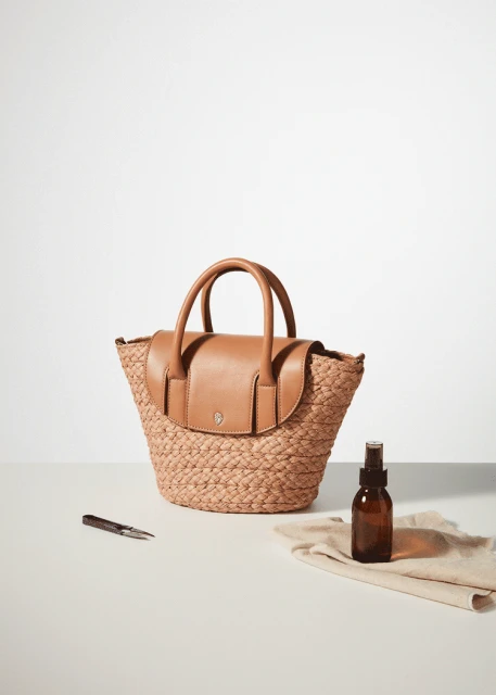 A straw tote bag with leather flap and handles sits on a light surface next to a brown spray bottle and cleaning cloth in a product shot.