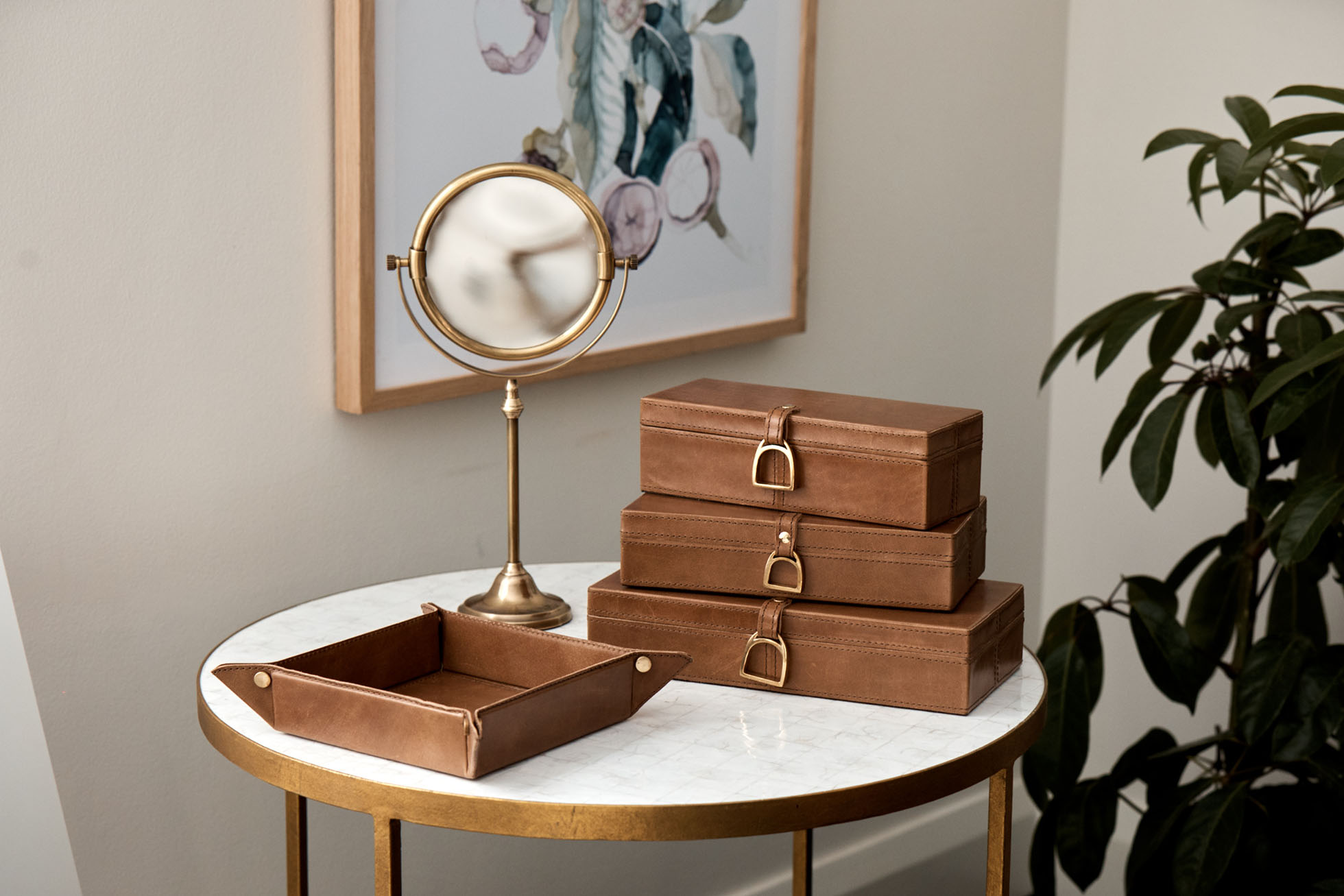 Stack of brown leather boxes with buckle closures and a gold-framed round mirror on a marble-topped table, showcasing home decor.