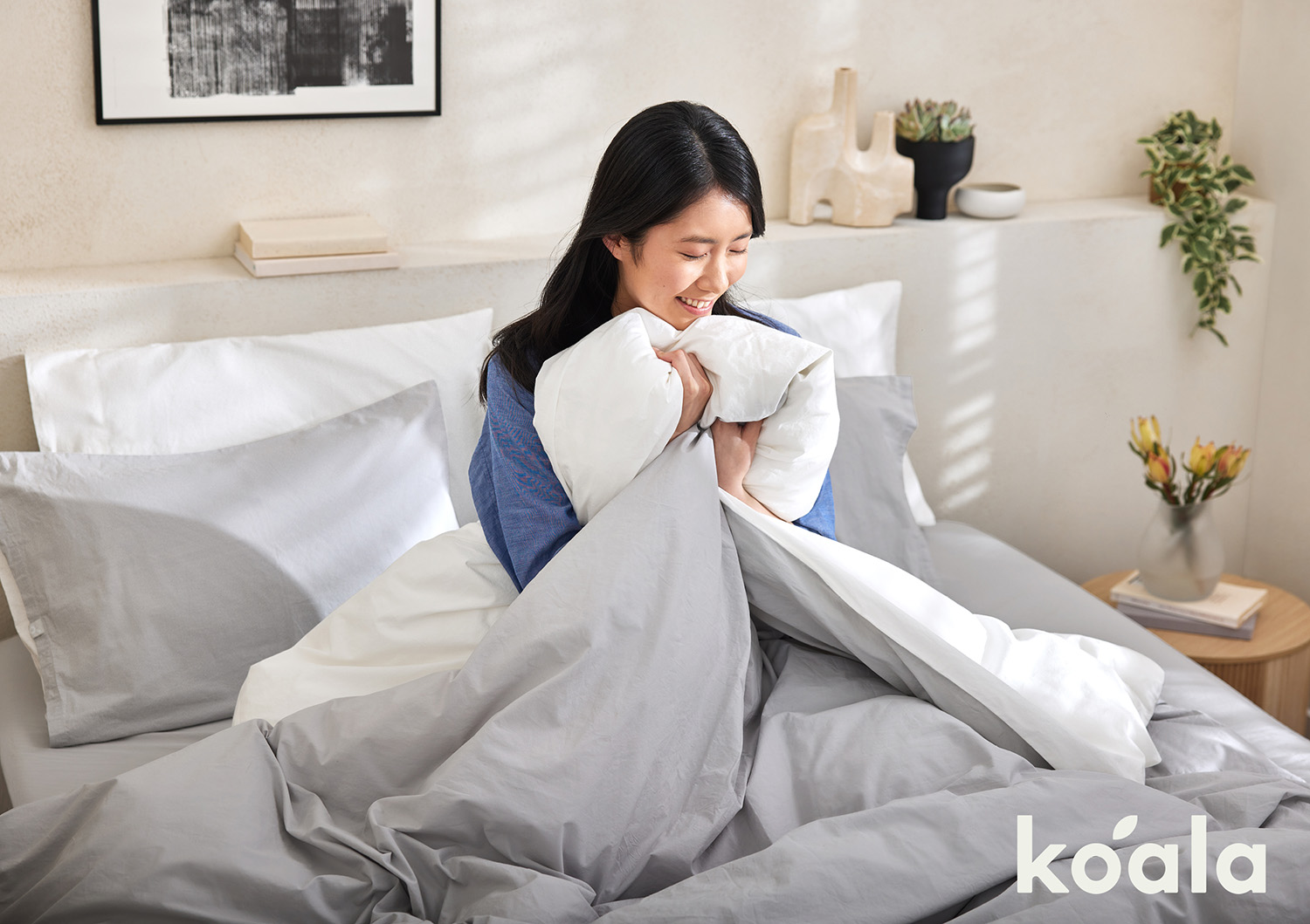 Smiling Asian woman sits in bed holding a white duvet against a cream wall with abstract art, plant, and soft natural lighting.