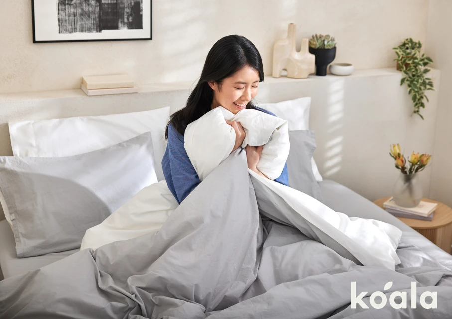 Smiling Asian woman sits in bed holding a white duvet against a cream wall with abstract art, plant, and soft natural lighting.