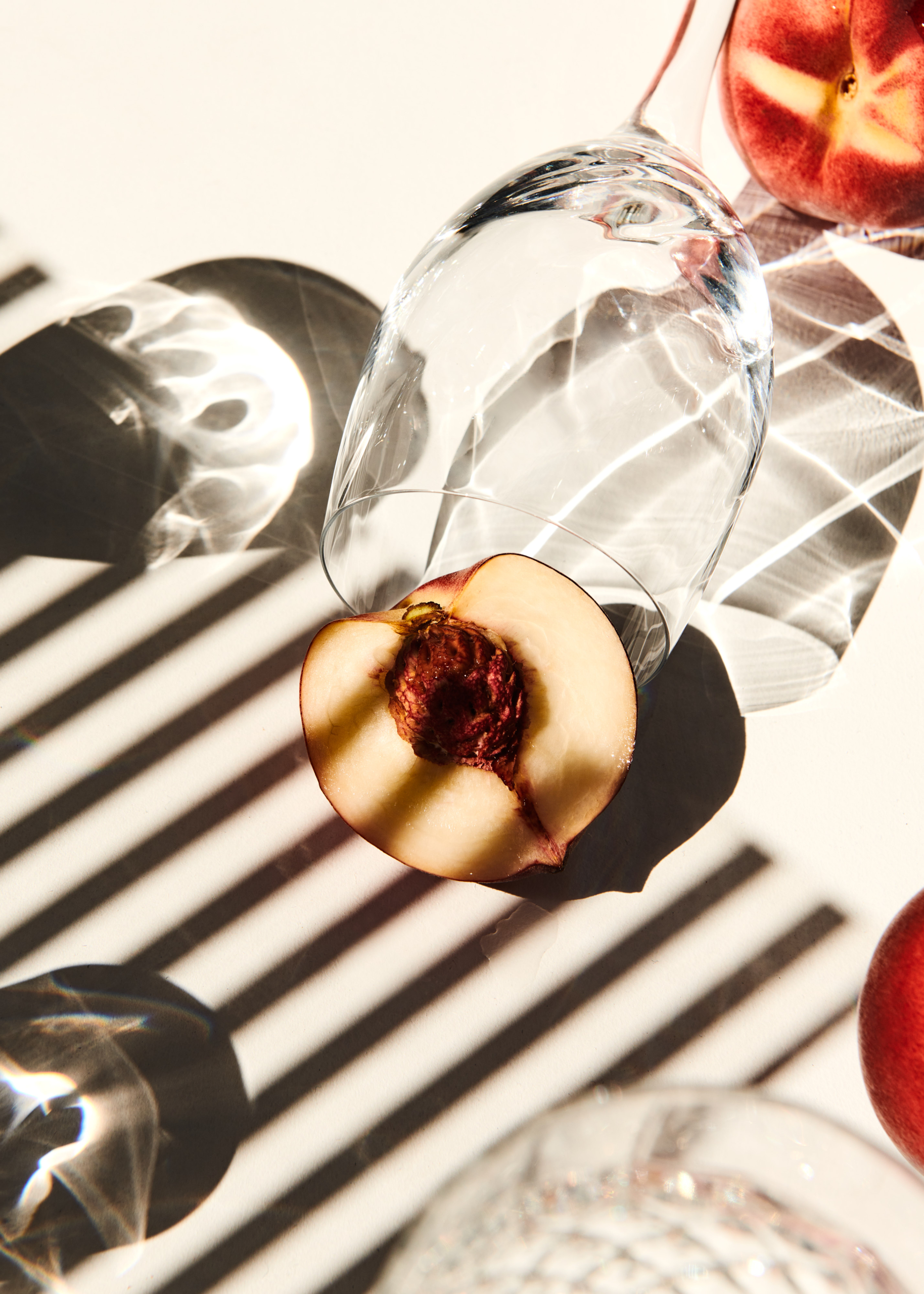Overhead shot of sliced peaches and overturned clear glasses on a white surface with striped shadows creating a modern, minimalist food still life.