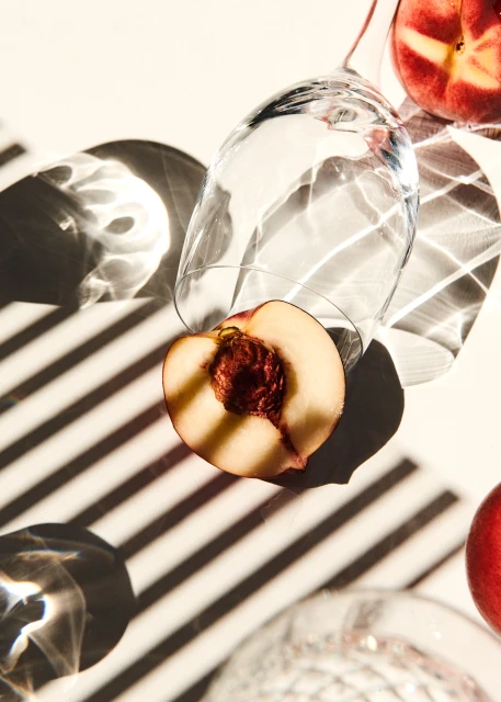 Overhead shot of sliced peaches and overturned clear glasses on a white surface with striped shadows creating a modern, minimalist food still life.