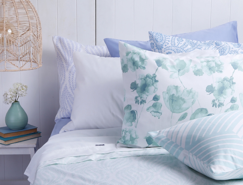 Bedroom decor with floral pillows on a bed, a woven pendant light, a vase of white flowers, and books on a bedside table.