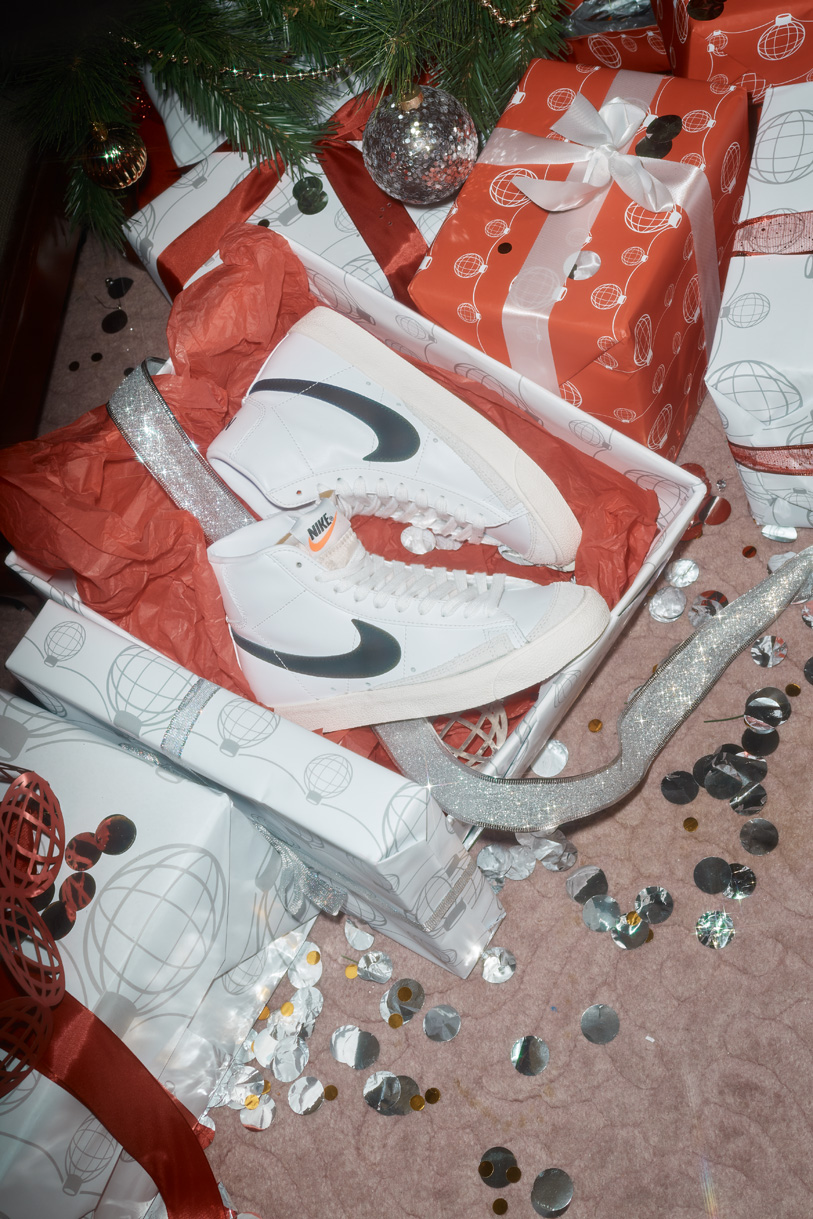 White high-top sneakers sit in a box with red tissue paper, surrounded by wrapped gifts, silver branch, and confetti; Christmas theme.
