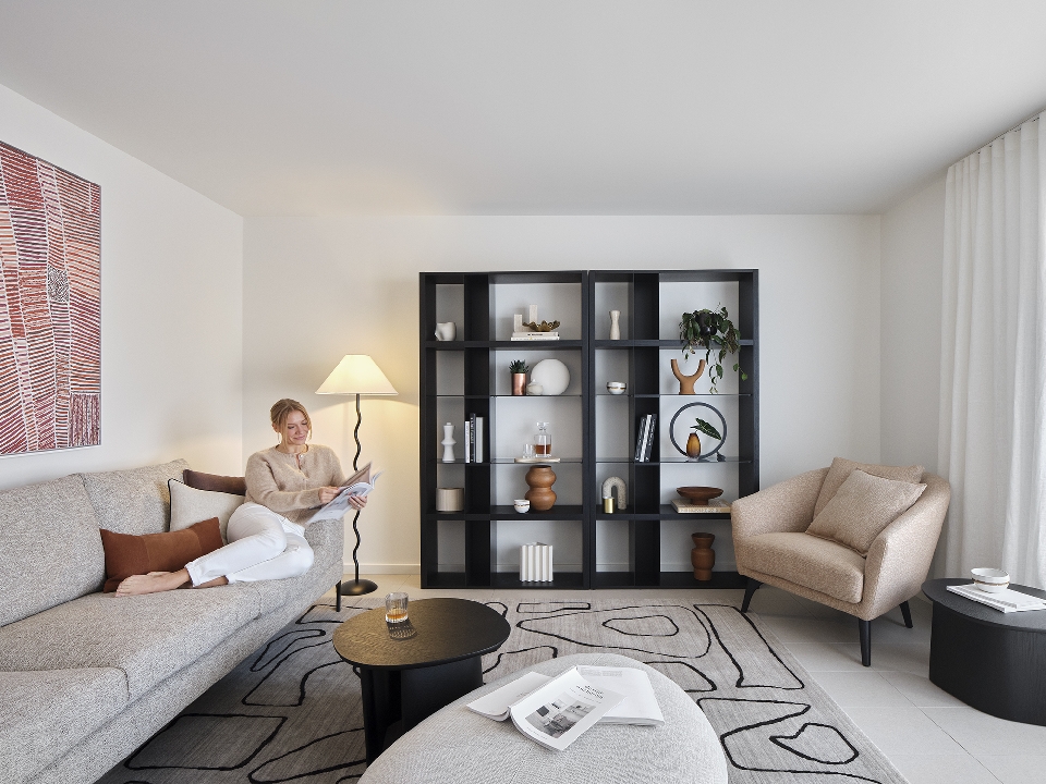 Woman reading on a couch in a living room with a black bookcase, tan armchair, oval coffee tables, and an area rug with line patterns.