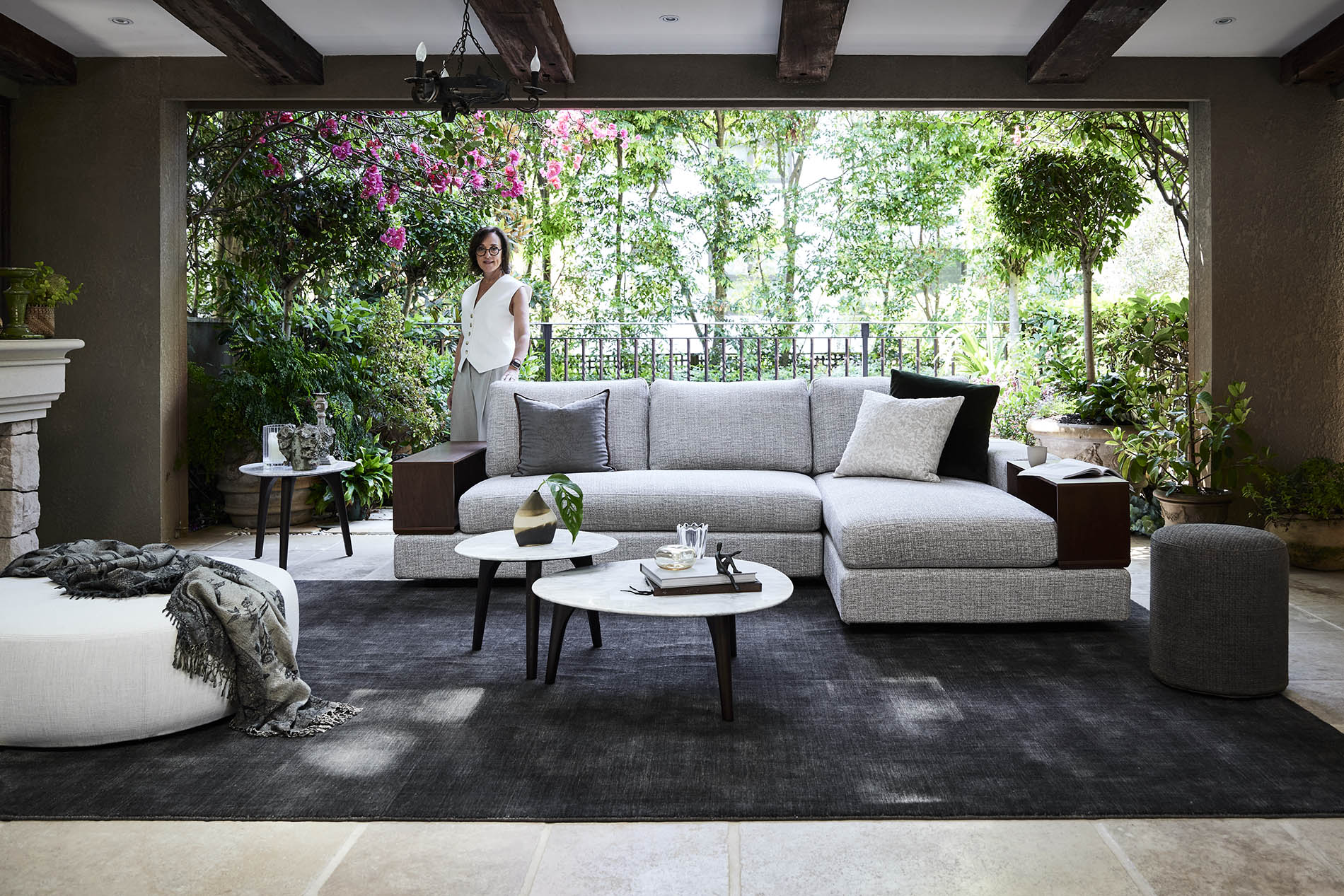 Living room interior features a grey sectional sofa, marble coffee tables, and a woman standing near lush greenery outside.