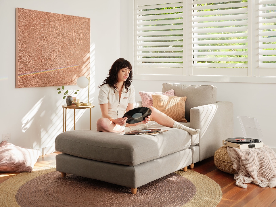 A young woman with dark hair sits cross-legged on an ottoman in a sunlit living room, holding a vinyl record, with neutral tones.