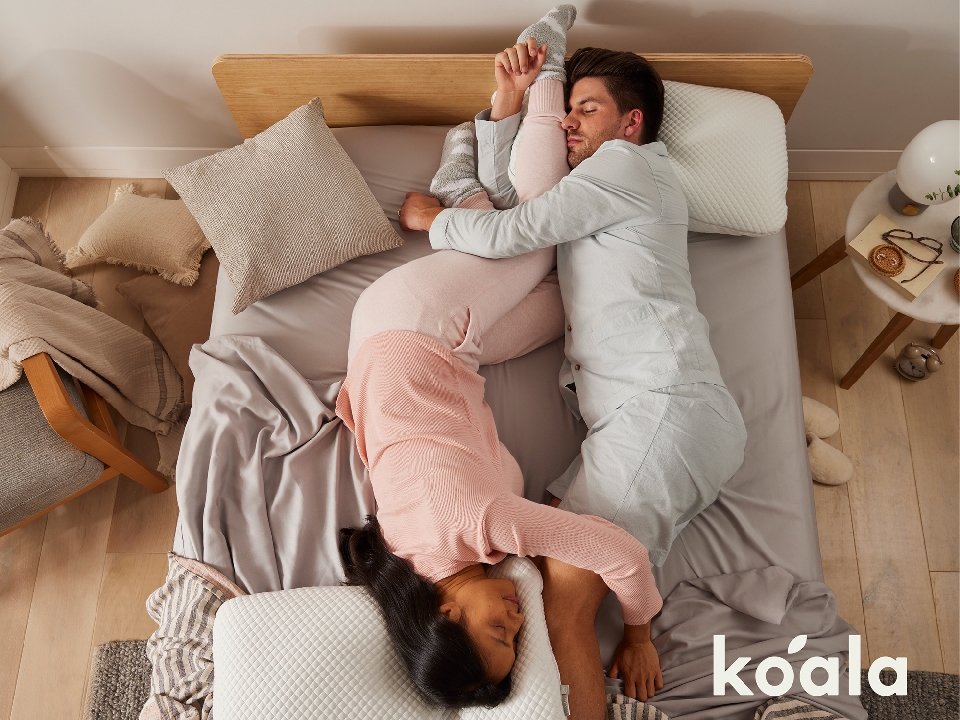 Overhead shot of a sleeping couple intertwined on a bed with pillows and bedding, with a logo in the bottom right corner.