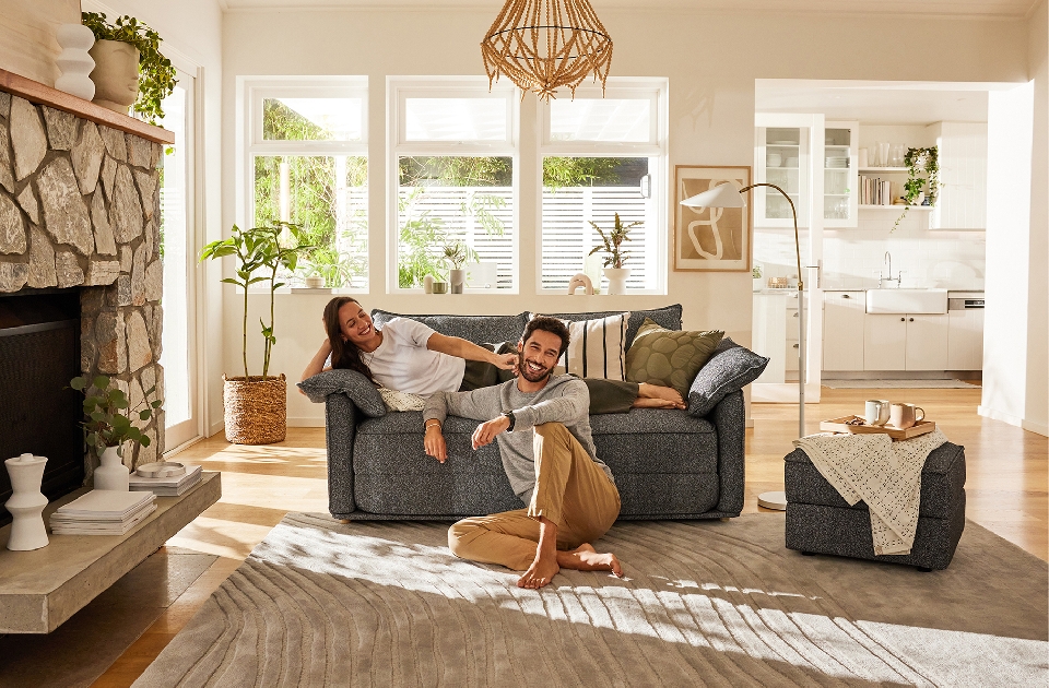 Living room interior with a young couple relaxing on a grey sofa near a stone fireplace and large windows, creating a warm home scene.