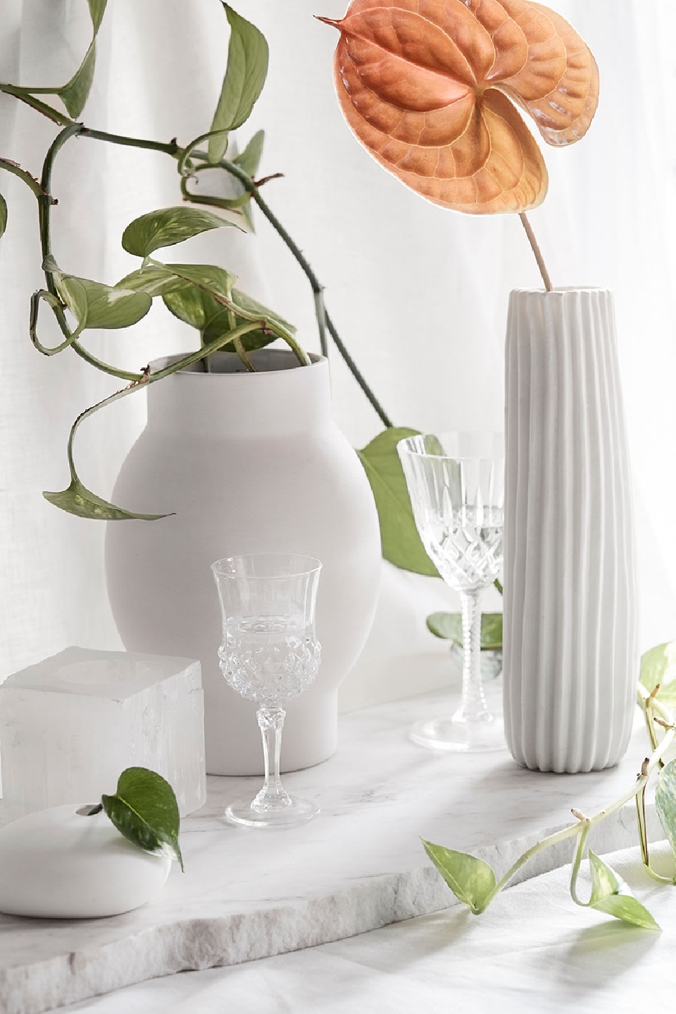 White and minimalist still life featuring assorted glassware, light-colored vases, and a marble surface with assorted plant life.