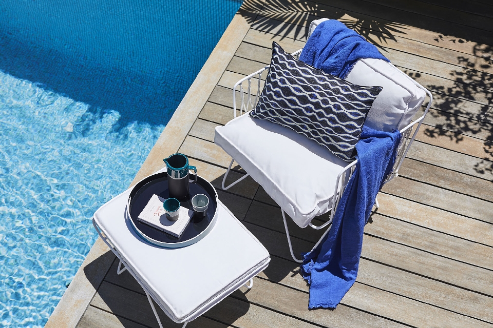 Outdoor poolside lounge area with a white wire chair, patterned cushion, blue towel, and a small table with a pitcher and glasses.