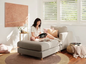 A woman seated on a grey chaise lounge holding a vinyl record, styled by Aurelie Tamin in a brightly lit interior space.
