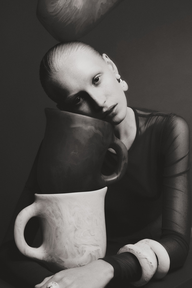 Monochromatic portrait of a bald woman leaning against pottery pitchers, featuring neutral tones and clay-like textures in an indoor setting.