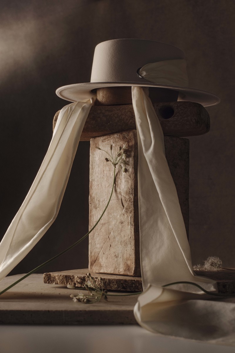 Still life of a beige hat with white ribbons on stacked wooden blocks, paired with delicate dried flowers against a gradient background.
