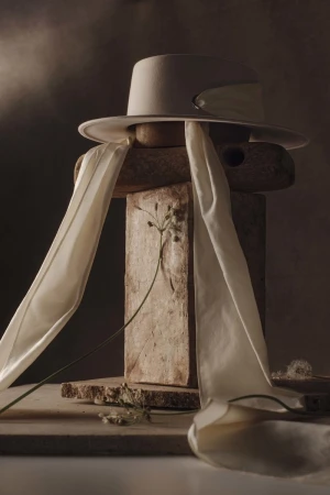 Still life of a beige hat with white ribbons on stacked wooden blocks, paired with delicate dried flowers against a gradient background.