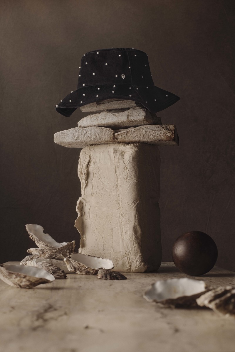 Still life of a black bucket hat with white polka dots balanced on stacked stones and oyster shells against a dark brown background.