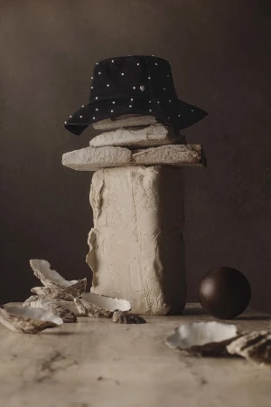 Still life of a black bucket hat with white polka dots balanced on stacked stones and oyster shells against a dark brown background.