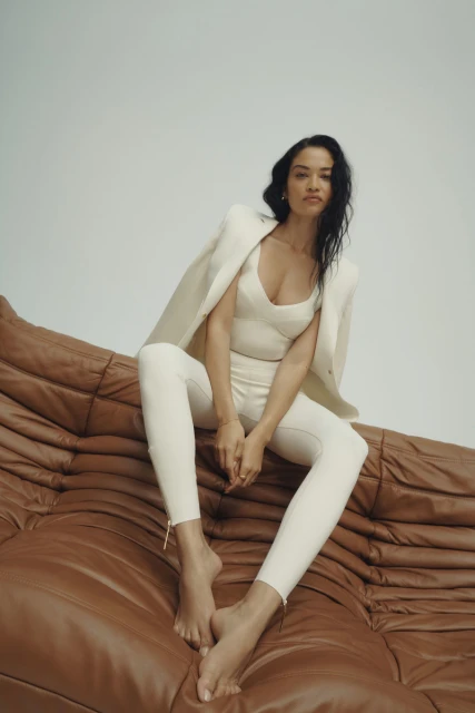 A woman in a cream-colored suit sits barefoot on a brown leather couch in a studio setting, looking directly at the camera with a neutral expression.