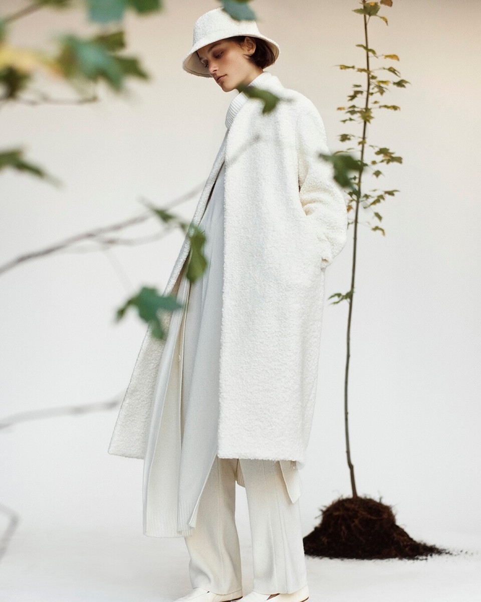 A woman wearing a long, white, textured coat and matching bucket hat stands against a white backdrop with tree branch accents.