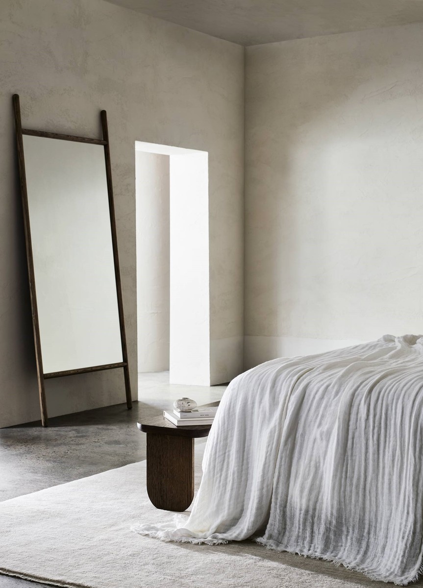 Minimalist bedroom interior with a leaning mirror, gray rug, wooden side table, and white throw blanket on a bed in neutral tones.
