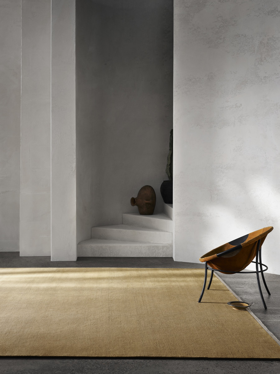Tan-colored rug on concrete floor beside modern chair in an indoor setting, with white walls and steps in the background. 