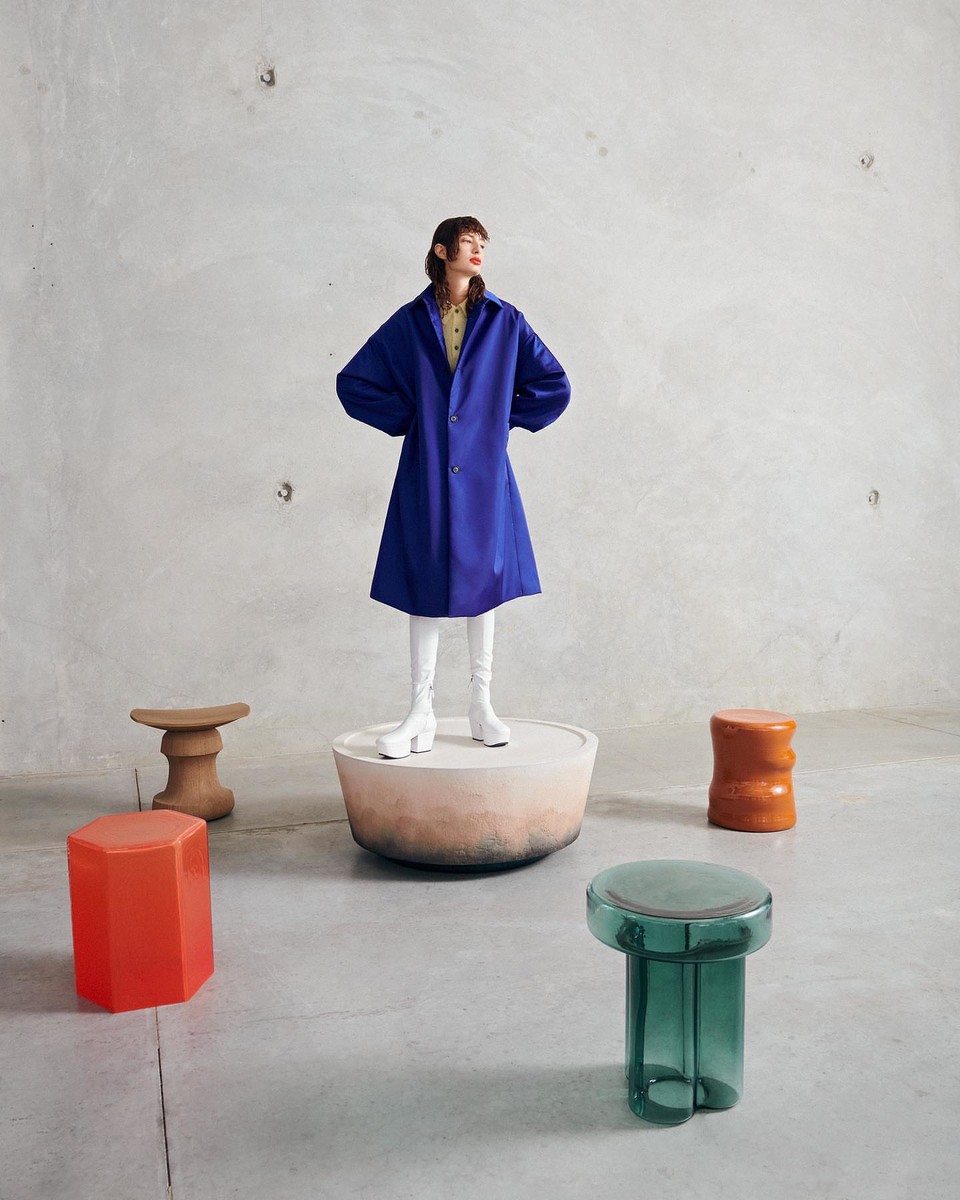 A woman stands on a concrete platform wearing a blue coat and white boots surrounded by modern stools in a neutral studio.