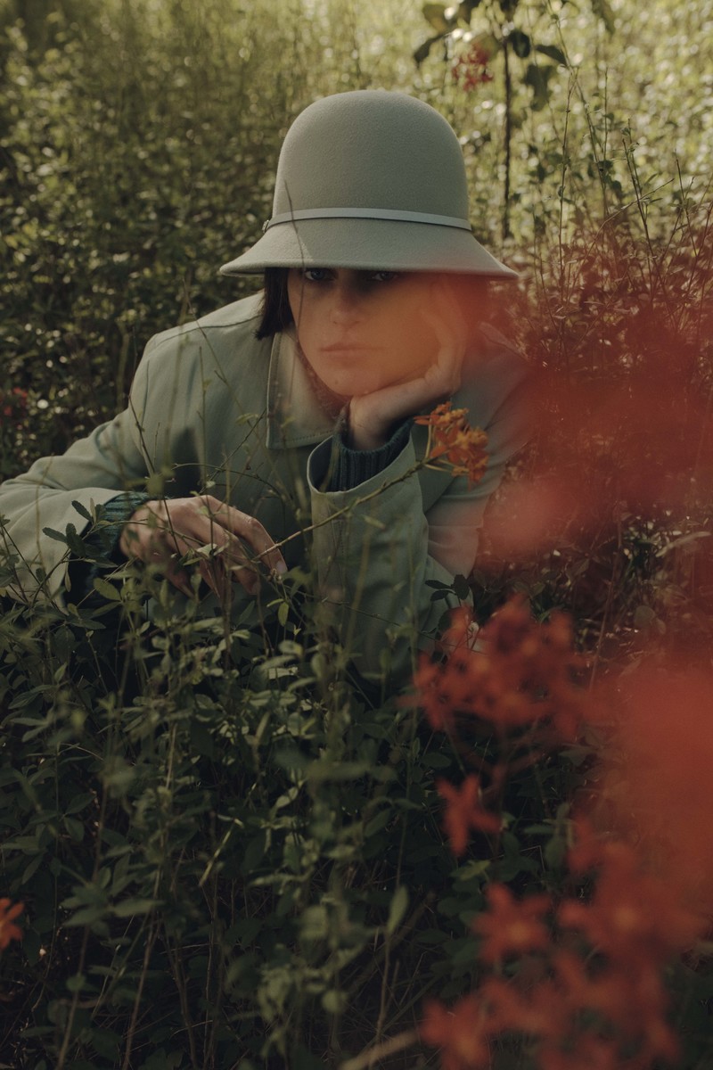 Woman in a bucket hat sitting amongst lush green plants and orange flowers. She is wearing a green jacket and looking off to the side.