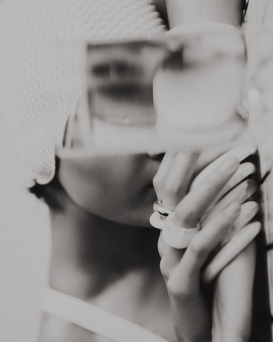Monochrome image featuring a woman's hands with rings, a hint of her face and a hat, and soft, diffused lighting.