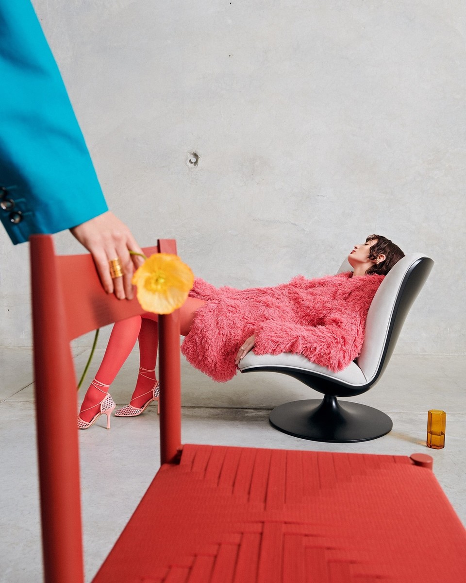 Fashion editorial shot featuring a person in a pink coat lounging in a chair, with another person holding a flower near a red chair.