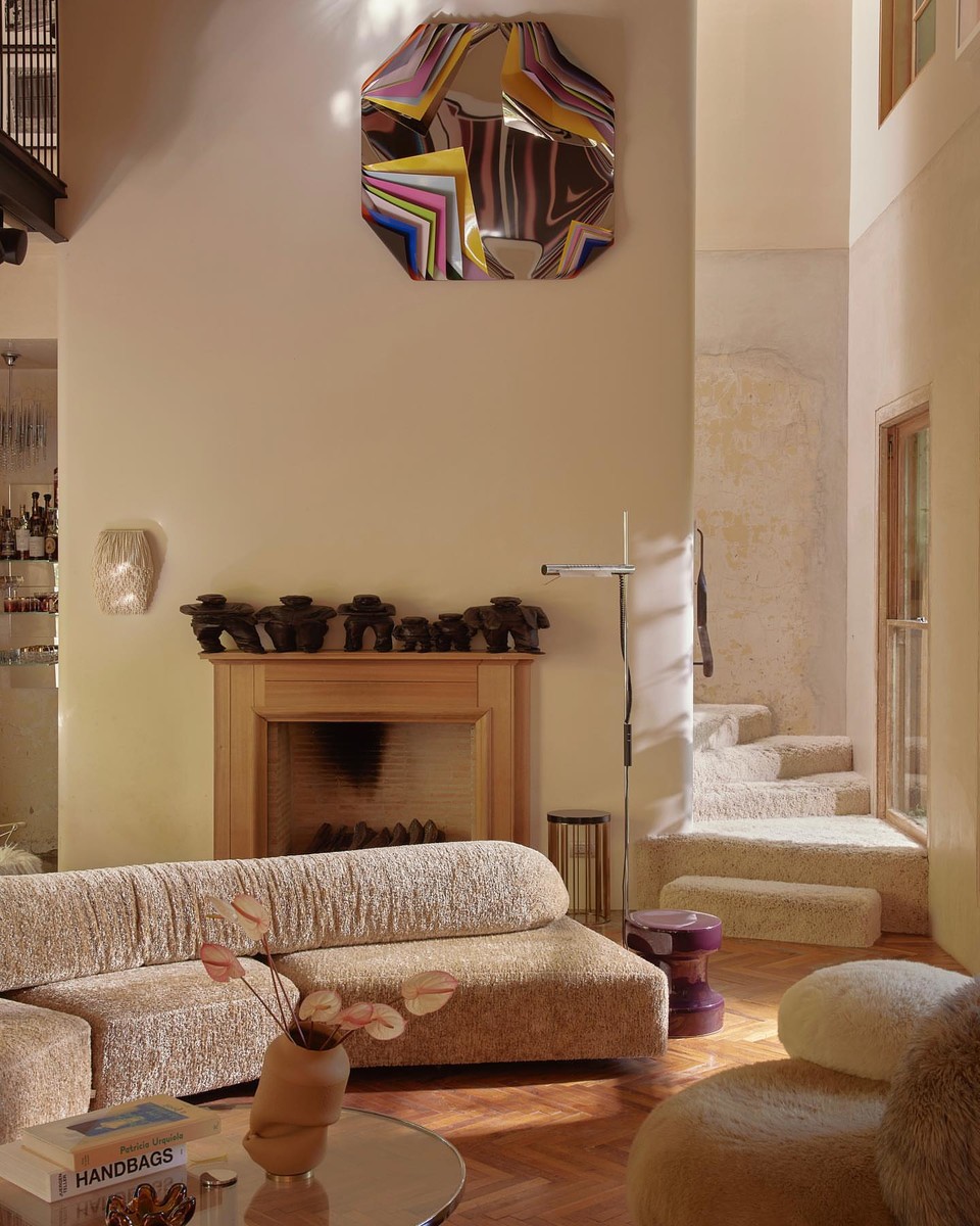 Living room interior with a modern sectional sofa, fireplace, and decorative mirror, illuminated by warm lighting and natural daylight.